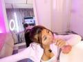 AlessiaFerry with brown hair in a ponytail, sucking her own finger, wearing a white top with cleavage, in a pink-lit room, reflected in a mirror.