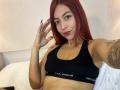 MiaaRouge with red hair, black sports bra, showing tits, tattoo on right arm, black nails, smiling, raising hand, sitting on bed, white pillow, red lipstick.