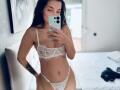 JuliCandy met donker haar, draagt een witte kant bh en slip, houdt een telefoon vast, toont een tatoeage op haar linkerarm, staat in een slaapkamer, en laat haar borsten en vulva zien..