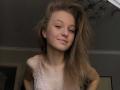 NievesMegee with long brown hair, slightly smiling, wearing a pink lace top, showing small tits, fair skin, and a nose ring, in a dimly lit room.