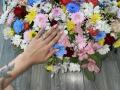 Olieri's hand with a tattoo, pink nails, and rings touches a colorful flower bouquet on a wooden floor. Blue, pink, red, and yellow flowers are spread around.