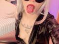 LaurenEvie69 with tongue out, black latex jacket, exposed tits, white hair, red lips, black sunglasses, tattoo on chest, in a bedroom. Her tongue is long and shiny.