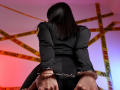 SkyeFrost with black hair, wearing a police uniform, cuffs on her wrists, chains, and hands behind her back, standing with "POLICE" tape in the background.