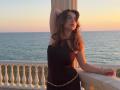 LeScharwath with wavy brown hair, black sleeveless top, gold chain, arm raised, standing by white railing, ocean sunset, hands on rail, looking off, soft evening light.