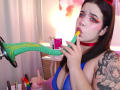 AryButty with green dildo in mouth, blue top, red choker, tattoo on arm, pink lipstick, rainbow sex toys, and a pink wall in the background.