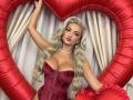 Mademoiselle Bella, com longos cabelos loiros, vestindo um corpete de veludo vermelho, exibindo seus seios grandes, segurando balões em formato de coração vermelho, um colar de pérolas, sexy, em pé em frente a cortinas brancas..