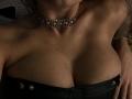 MademoiselleBella's tits are visible in a black strapless top, wearing a silver chain necklace, with her hands on her chest, in dim, moody lighting.