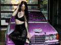 AlexLovely with long black hair, yellow glasses, black bra, lace pants, tattoos, and dark lipstick, sitting on a purple, leopard-print, soapy car, hand in hair, looking sexy.