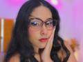 MarilynHills with long black hair, glasses, and red lipstick, touching her cheek with a hand, has a serious expression and is wearing a black lace top.