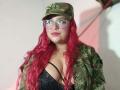 Estefy69 with red wavy hair, glasses, camo cap, and camo jacket, showing big tits in a black lace bra, smiling. She's chubby with pink lipstick.