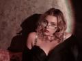 AdrianePittillo with wavy blonde hair, green glasses, black off-shoulder dress, showing her tits, dark shadow, pink lace, and a black garment on her right.