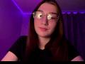 SunnyKlusman with glasses, brown hair, and nose ring, wearing a black shirt, smiling slightly, in a purple-lit room.