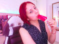 StephanieSun with red hair, eating a pink popsicle, wearing a black sleeveless dress with white patterns, sitting in a chair, looking at the camera, holding the popsicle in her mouth.