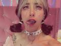 LaurenEvie69 with teary eyes, tongue out, drooling, wearing a spiked collar, pigtails, and a flower tattoo on her shoulder, in a pink, tufted, headboard.