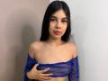 NaomyRosse with long black hair, wearing a sheer blue top, covering her tits with one hand, looking at the camera with red lipstick, standing against a plain wall.