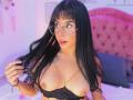 GraceClarke with black hair, glasses, and a gold chain top, exposing her big, round, bare tits. She's holding her hair, looking at the camera with pink lips.