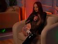 TorriKitt with red hair, wearing black lace bodysuit, exposing her tits, and black boots, sitting on a beige chair, showcasing a large thigh tattoo, in a dimly lit room.