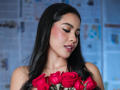 PaolaYKatty with dark, wet-looking hair, closed eyes, and pink lipstick, holding a bouquet of red roses, showing off her shiny, blushed cheeks and upper body.