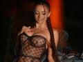 AishaBee with long braided hair, wearing a black mesh top that reveals her big boobs, a gold necklace, and red nails, standing next to a motorcycle.
