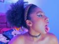 MeganWlliams with dark curly hair in a leopard print headband, tongue out, wearing bold eyeliner and a black choker, blue and purple lighting on her bare shoulders.