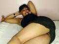 RareGem, lying on a bed, wearing a black dress, has her tongue out, arms behind her head, exposing her ass and pussy, with a black bracelet on her wrist.