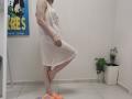 JoannaSia standing in white lace dress, one leg raised, showing bare ass, with orange slippers on tiled floor, potted plant in corner, and "ZERTS" poster on wall.