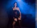 AgataRyili with dark lace corset, black garter stockings, and chain, standing in blue fog, gripping a chain, shadowed by striped light, showing her tits and ass.