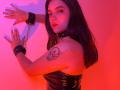 Katarina Novak met zwarte leren manchetten en een ketting, roze-rode verlichting, zwart haar, kijkt naar de camera, tatoeage op de rechter schouder, zwart leren top zonder bandjes, handen tegen de muur..