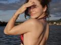PeacHub69 with wet skin, smiling, covering eyes, in a red and black bikini top, arm raised, wearing a bracelet, standing in water, sunlight on back, brown hair.