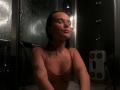 EsterGillom in a shower, wet with water droplets, wearing a thin, pink, bra-like top, eyes closed, black tattoos on arms, dark wet hair, slight smile, water streaming.