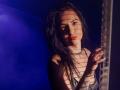 IngaDrein with wet black hair, red lipstick, and a chain necklace, standing in shadow with striped light patterns on her face, holding a fence with blue light.