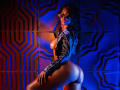 Kinkyandsavage, avec une peau foncée, des cheveux bouclés, portant des lunettes de soleil et une veste en satin bleu, exposant ses seins et ses fesses, agenouillée, éclairée par des lumières bleues et oranges..