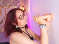 StephanieSun with red hair, glasses, and black choker, sucking on a large, flesh-colored dildo, her hands gripping it, wearing a silver watch and black bracelet.