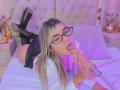 SashaPolet, with glasses and blonde hair, is on a bed, sucking a cock, wearing black boots and a white shirt, with a microphone in the foreground.