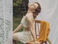 TeresaBirt with glasses, wavy brown hair, floral off-shoulder top and skirt, sitting on wooden chair, yellow blanket, white lace curtain, head tilted back, eyes closed, looking relaxed.