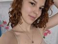 ChayaParrett has curly red hair, light skin, and small tits. She's wearing a beige tank top and a necklace with a small pendant. She's smiling slightly with a hand in her hair.