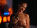 RosaInk, topless, with dark curly hair, tattoos on arms, holding her tits with tattooed hands, wearing a pearl necklace, and a red bracelet, smiling softly.