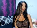 BrunetteBabe69 with long black hair, wearing a black mesh top, exposing her big boobs, looking at the camera with a pouty expression, in a dimly lit room.