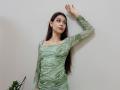 AmeliaMaiv with long brown hair, wearing a green marbled dress, standing against a wall, arm raised, looking to the side, small earrings, pouty lips, plant in the bottom left.
