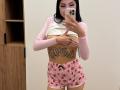 AuraXo with long black hair, pink long-sleeve shirt, white crop top, strawberry-patterned pink shorts, showing a tattoo on her flat stomach, holding a phone in a mirror selfie.