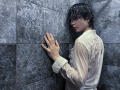 RubyMote with wet black hair, wearing a transparent white shirt, touching gray tiled wall with right hand, looking over shoulder, in a dimly lit shower.