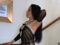 Vivienne West con lunghi capelli neri, che alza un braccio, indossa un abito nero trasparente con spalline bianche, che rivela l'ascella, in piedi vicino a una scala, guarda di lato, rossetto rosso..