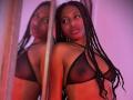 QueenPerlaa with long black braids, wearing a sheer black bra, showing small nipples and a soft smile. Her reflection is in the mirror, highlighting her dark skin.