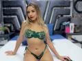 CloeRouss is sitting on a bed, wearing green lace lingerie with her tits and pussy covered, blonde hair, tattoos, red lipstick, and a necklace, looking at the camera.