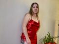 Aminas in a red satin dress, showing her tits and a tattoo on her right thigh, standing with a pensive look, next to a green plant.