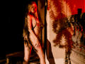 MiaMeowa with long blonde hair, naked, wearing a chain around her tits, standing in shadowy red light, hands on her ass, next to a stone column, photo.