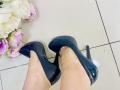 Olivia Grandes glänzende schwarze Stilettos auf ihren blassen Füßen, mit rosa und weißen Blumen auf der linken Seite. Fliesenboden im Hintergrund. Ihre Zehen sind leicht nach innen gekrümmt im Inneren der Schuhe..