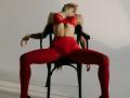 MilagrosEdmisten with red lace bra, red pantyhose, and black chair, head tilted back, arms resting, legs spread, exposing small tits, red panties visible, in a provocative pose.