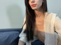 Valnes with long black hair, pink lips, and light skin, wearing a sheer white top, exposing her small tits and bare chest. She's sitting on a blue couch.