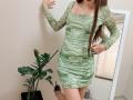 AmeliaMaiv wearing a green, marbled dress with long sleeves, standing with one hand on a wall, her long brown hair down, black heels, and a plant in the background.